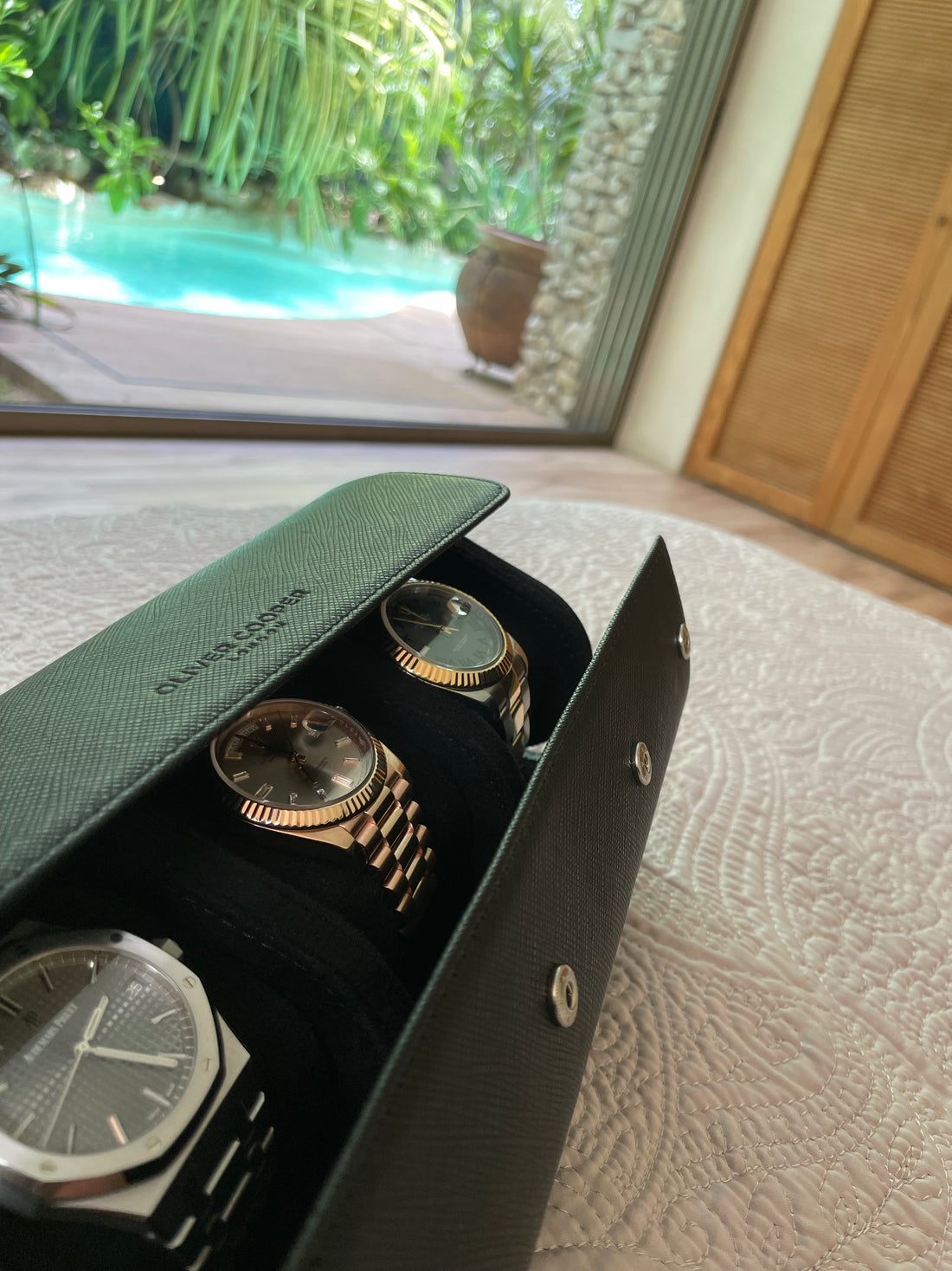 Black leather watch roll with three luxury watches inside, resting on a bed in a modern bedroom with a swimming pool in the background.