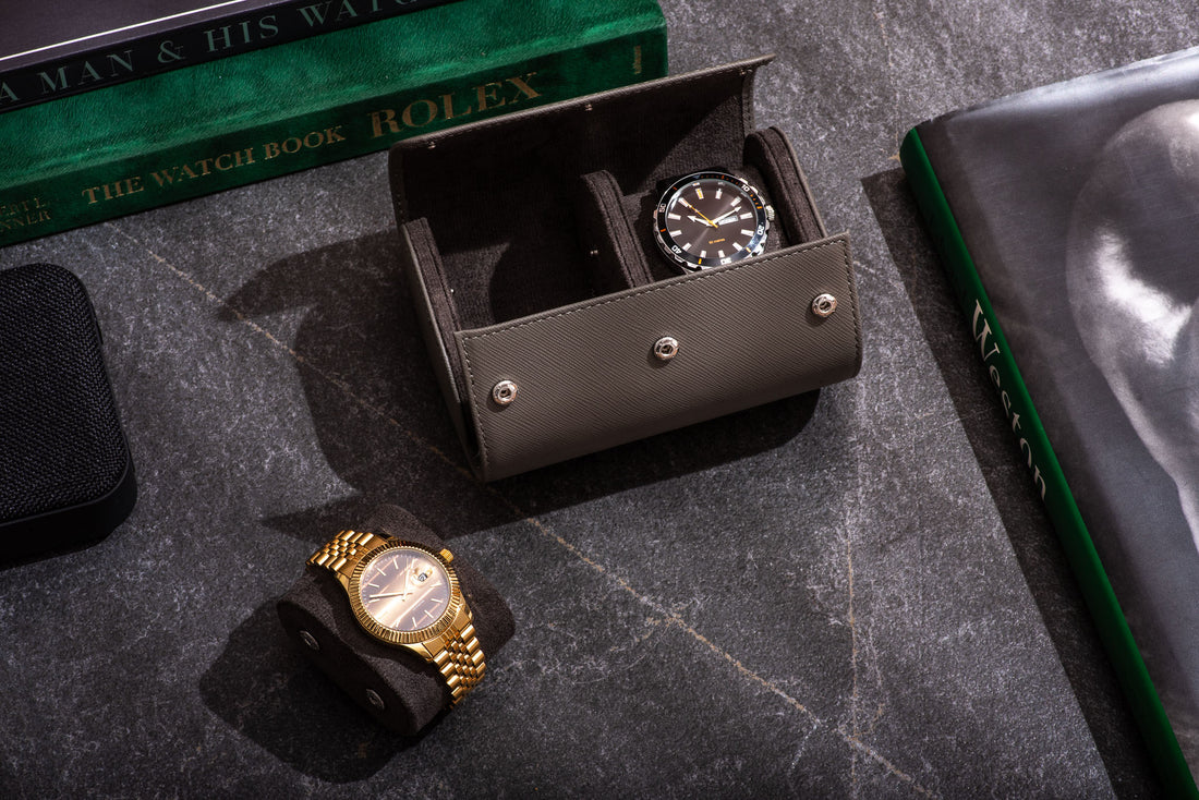  Two gold colored wristwatches with black leather bands on a wooden table next to a closed, brown leather watch case with gold text