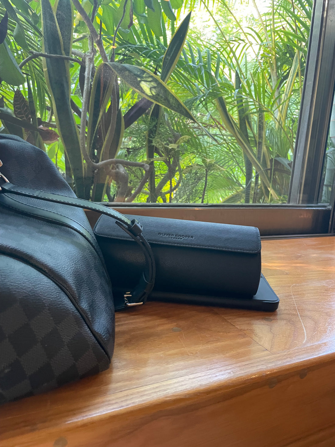   A brown leather purse with a gold zipper closure sitting on a wooden table next to a black glasses case with a gold Oliver Cooper logo. 