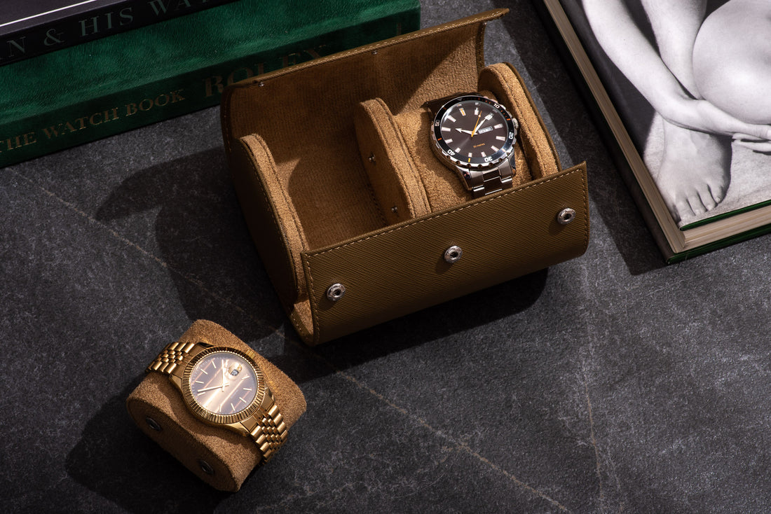  Two gold colored wristwatches with black leather bands displayed on a wooden table. A closed, brown leather watch case with gold text sits beside the watches.