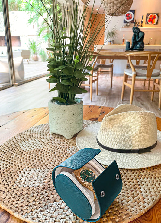  A brown hat and a silver wristwatch sit on a wooden table with a potted plant in the background.