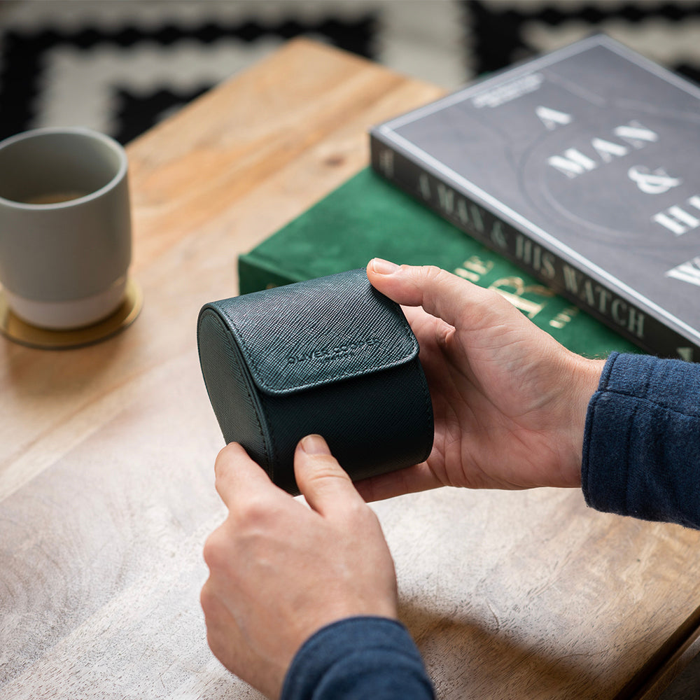 Man holding a single watch case with a real leather exterior, surrounded by watch books and a coffee mug on a wooden table.